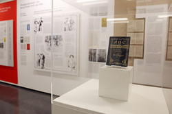 Ausstellungsansicht aus ‚Benehmt euch‘ im Dortmunder U: Eine Glasvitrine mit einem Schriftstück vor Schrift- und Bildtafeln an den Wänden.