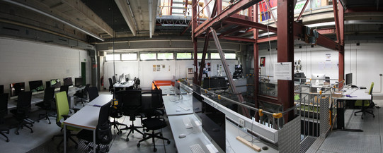 Panoramaansicht eines Arbeitsraums in den Kunsthallen der TU Dortmund mit Mac-Pool: Zahlreiche Computerarbeitsplätze mit Monitoren und schwarzen Bürostühlen stehen entlang der Wände und auf langen Arbeitstischen. Im Hintergrund befinden sich Fenster mit Tageslicht, im Rauminneren ragen Stahlträger und technisches Equipment in die Höhe. Die industrielle Architektur mit offenen Decken und roten Stahlträgern verleiht dem Raum einen modernen Werkstattcharakter.