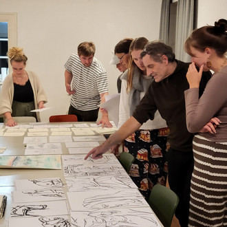 Studierende und Lehrende stehen um einen Tisch auf dem Zeichnungen liegen
