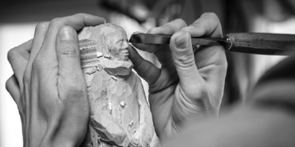 Zwei Hände halten ein Stück Holz aus dem sie eine magere Figur schnitzen. Fotografie in schwarz-weiß