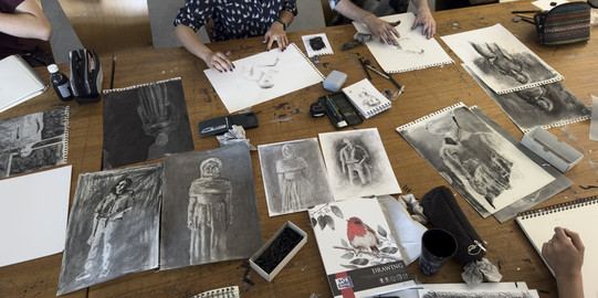 Studierende und Lehrende sitzen um einen Tisch auf dem Zeichnungen liegen