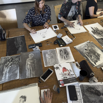 Studierende und Lehrende sitzen um einen Tisch auf dem Zeichnungen liegen