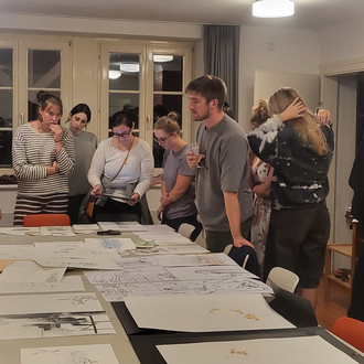 Studierende und Lehrende stehen um einen Tisch auf dem Zeichnungen liegen