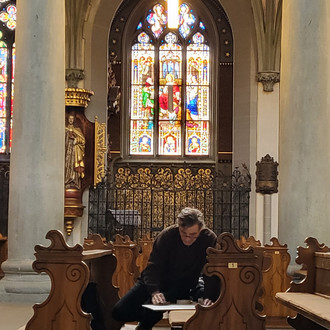 zeichnende Person in einer Kirchenbank vor buntem Kirchenfenster im Innenraum einer Kirche.