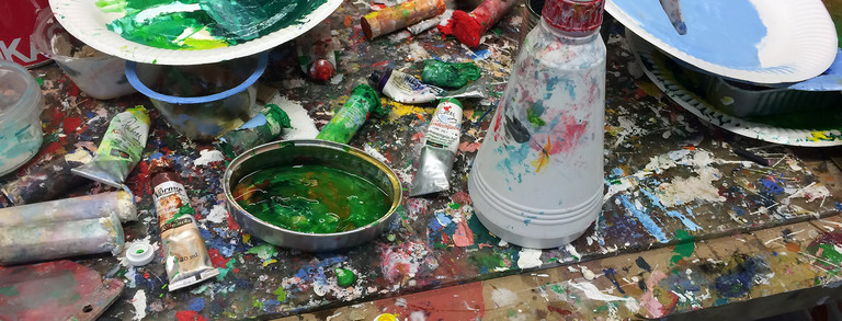 Ein stark genutzter Arbeitstisch in einem Atelier: Auf der mit Farbspritzern bedeckten Tischplatte liegen zahlreiche Farbtuben, Pinsel, Deckel mit Farbe, Pappteller mit grüner und blauer Farbe, ein Farbsprühbehälter sowie verschiedene Malutensilien. Unter dem Tisch sind weitere Farbtuben sichtbar. Das Bild vermittelt einen Eindruck von intensivem künstlerischem Arbeiten.