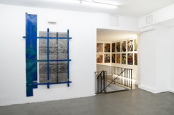 Ausstellungsansicht im Superraum Dortmund: links ein wandmontiertes Werk mit blauen, gitterartigen Elementen, rechts Blick in einen angrenzenden Raum mit dicht gehängten, kleinformatigen Portrait-Gemälden, Ausstellung TPLI III.