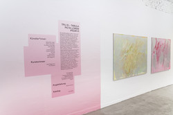 Seitliche Perspektive im Ausstellungsraum Superraum Dortmund: rosa Wandtext mit Informationen zur Ausstellung TPLI III, rechts zwei abstrakte Gemälde in Gelb- und Rosatönen an weißer Wand.