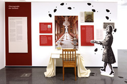 Ausstellungsansicht aus ‚Benehmt euch‘ im Dortmunder U: Ein gedeckter Tisch mit Stuhl steht vor einer großen Fotografie eines prunkvollen, festlich gedeckten Saals. Umgeben ist die Szene von roten Texttafeln und historischen Abbildungen. Rechts daneben befindet sich eine große schwarz-weiße Figur eines Dieners mit Tablett