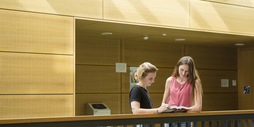 Zwei Studentinnen stehen mit einem Buch am Geländer.