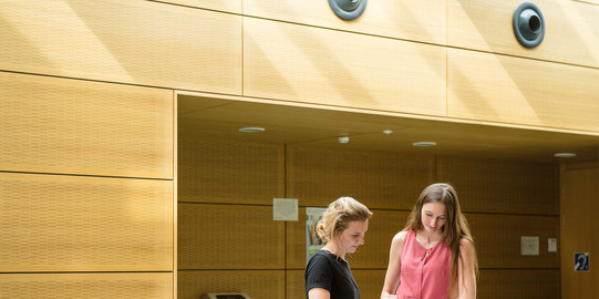 Zwei Studentinnen stehen mit einem Buch am Geländer.